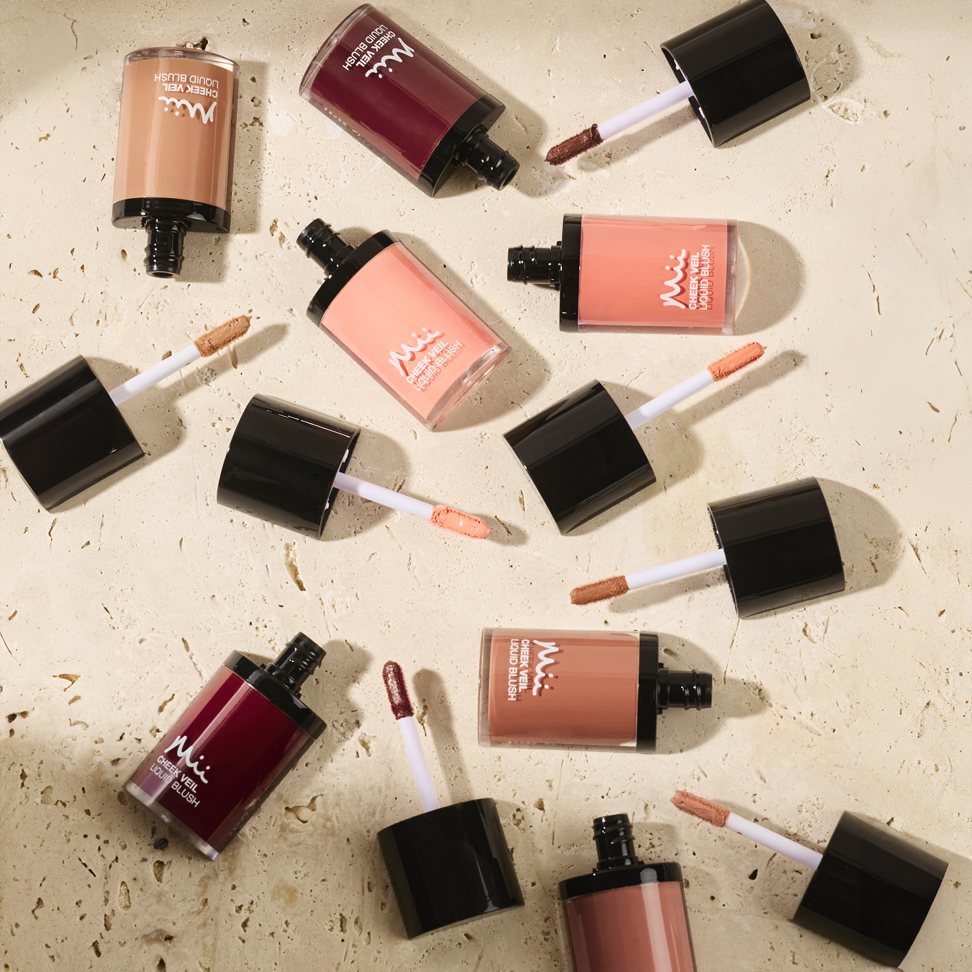 Assorted shades of Mii Cosmetics Cheek Veil Liquid Blush bottles with applicator wands displayed on a beige stone surface.